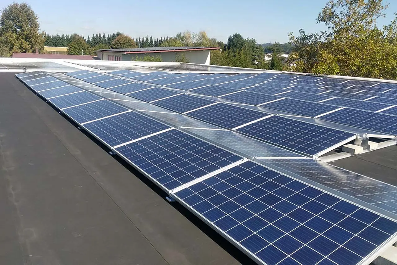 Aufgeständerte Photovoltaikmodule auf einem Flachdach, dahinter Gewerbegebäude und bewaldete Hügel